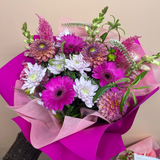A vibrant waterbox featuring magenta and white gerbera daisies, pink chrysanthemums, and green foliage, wrapped in bright pink tissue paper and pink paper wrap, set against a solid beige background.