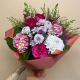 A waterbox of mixed flowers including pink hydrangeas, magenta roses, white and purple chrysanthemums, and green foliage, wrapped in dusty pink paper against a plain beige background.