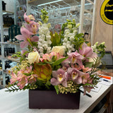 A vibrant floral arrangement in a dark rectangular planter box, featuring pink orchids, white roses, and green foliage, displayed on a white counter inside a shop.