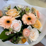 A bouquet of peach and apricot  flowers wrapped in white tissue paper and a sheer beige wrapper, featuring peach and cream roses, gerbera daisies, and green foliage.