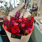 A vibrant mixed flower bouquet held by a hand inside a flower shop. The bouquet features red, pink, and purple flowers wrapped in white paper with red tissue paper and a black label.