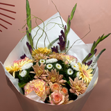 A mixed flower bouquet wrapped in white tissue paper and a light beige paper sleeve, featuring peach roses, yellow and white daisies, peach dahlias, and green foliage, set against a solid dusty pink background.