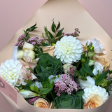A wrapped flower bouquet featuring white dahlias, peach roses, green succulents, and small pink flowers, wrapped in beige paper against a soft pink background.