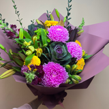 A vibrant mixed flower bouquet held by a hand wearing a light pink sleeveless top. The bouquet features prominent pink chrysanthemums, yellow daisies, green foliage, and dark purple ornamental kale, wrapped in a dusty purple paper wrapper.